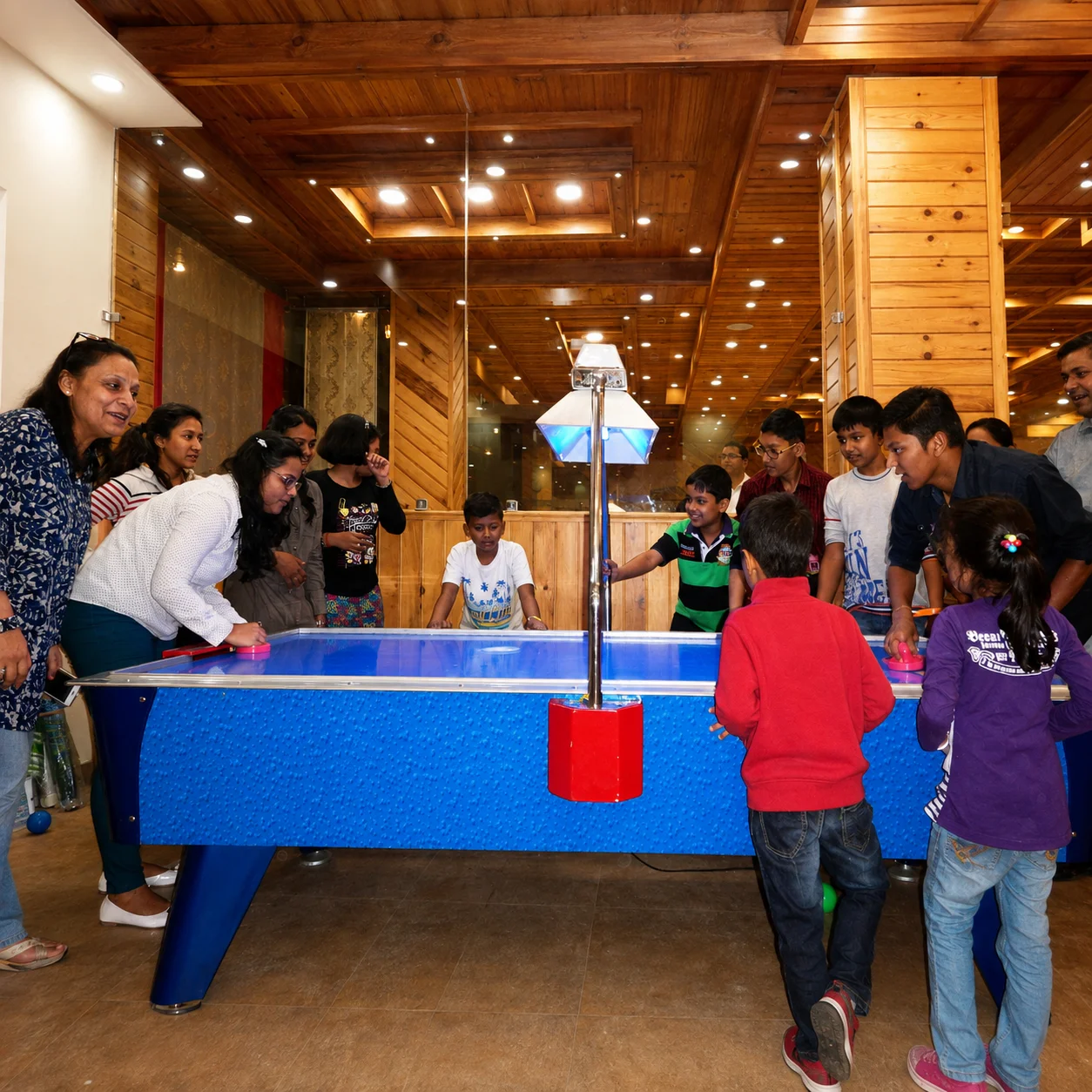 Children and adults gathered around a blue air hockey table in a wood-paneled game room.